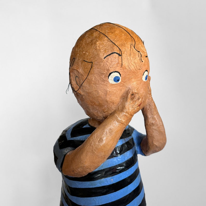 Puede incluir: Una escultura de papel mach&eacute; de un ni&ntilde;o con ojos azules y cabello casta&ntilde;o que lleva una camisa a rayas azules y negras. El ni&ntilde;o se tapa la cara con las manos.
