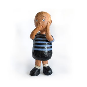 Puede incluir: Una figura de papel mach&eacute; de un ni&ntilde;o con ojos azules, que lleva una camisa a rayas azules y negras y pantalones negros. Se est&aacute; tapando la boca con la mano y tiene una expresi&oacute;n de sorpresa.