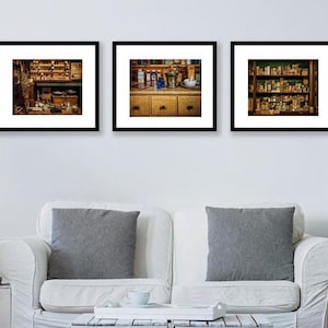 May include: Three framed prints of an apothecary shop interior. The prints depict shelves of bottles, drawers, and various tools. The prints are displayed above a white couch with gray pillows. A small side table with a plant and books is next to the couch.