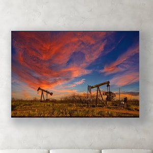 May include: A framed canvas print featuring two oil pump jacks silhouetted against a vibrant sunset sky. The sky is a mix of orange, red, and blue hues, with the pump jacks set in a field of dry grass.
