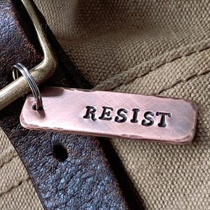 May include: A copper-colored metal tag with the word "RESIST" engraved on it. The tag is attached to a brown leather strap with a brass buckle.