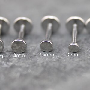May include: Five silver-toned metal labret studs with flat circular tops arranged in a row. The sizes are labeled below each stud as 3.5mm, 3mm, 2.5mm, 2mm, and 1.5mm, indicating the diameter of the flat circular top of each individual stud.