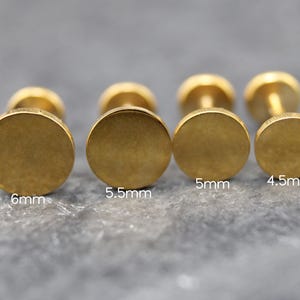 May include: Four gold-colored stud earrings with flat circular faces arranged in a row. The sizes are labeled as 6mm, 5.5mm, 5mm, and 4.5mm, indicating the diameter of each circular face. The earrings have a smooth, matte metallic finish.