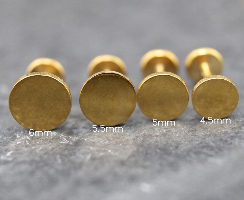 May include: Four gold-colored stud earrings with flat circular faces arranged in a row. The sizes are labeled as 6mm, 5.5mm, 5mm, and 4.5mm, indicating the diameter of each circular face. The earrings have a smooth, matte metallic finish.