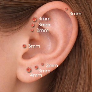 May include: An ear with multiple orange gemstone stud earrings. The sizes are labeled as 2mm, 3mm, and 4mm. The earrings are set in a silver or white metal. The ear is shown against a blurred background.
