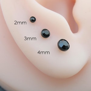 Può includere: Primo piano di tre orecchini a bottone neri di diverse dimensioni (2 mm, 3 mm e 4 mm) su un orecchio. Gli orecchini hanno un aspetto sfaccettato, simile a una gemma. Le dimensioni sono indicate in testo nero.