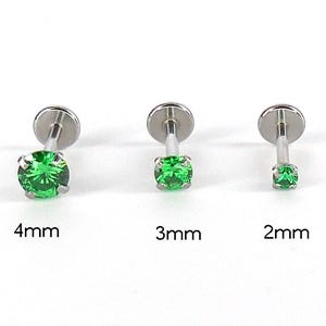 May include: Three silver-colored labret studs with green gemstones. The sizes are 4mm, 3mm, and 2mm. The gemstones are round and faceted, and the posts have a flat disc on the back.