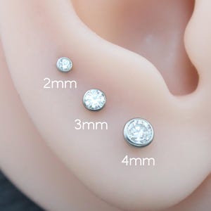 Può includere: Tre orecchini a lobo rotondi con pietre trasparenti sfaccettate incastonate in metallo color argento. Gli orecchini sono disposti in fila, etichettati con le misure 2mm, 3mm e 4mm, mostrando la scala di ogni pezzo in base al diametro.