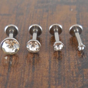 May include: Four silver barbell earrings with clear crystal gems. The earrings are different sizes, with the smallest on the right and the largest on the left.