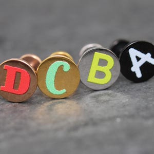 May include: Four circular stud earrings arranged in a row. The first is rose gold with a red letter D, the second is gold with a mint green letter C, the third is silver with a neon yellow letter B, and the fourth is black with a white letter A.