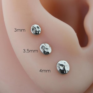 Può includere: Primo piano di tre orecchini a bottone argentati con texture martellata. Gli orecchini sono di 3 mm, 3,5 mm e 4 mm. Gli orecchini sono esposti su un orecchio rosa chiaro. Gli orecchini hanno un aspetto strutturato e martellato.