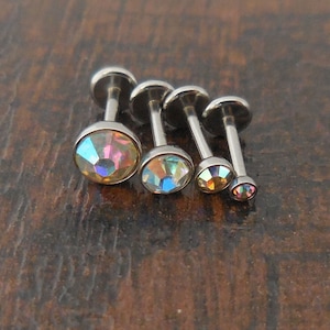 May include: Four silver body jewelry pieces with iridescent gemstones. The gemstones are different sizes and are set in a round shape. The jewelry is made of surgical steel.