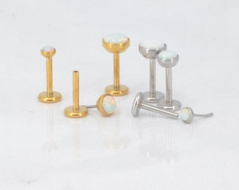 May include: An assortment of labret studs with opal-like cabochon tops. Some studs have gold-colored posts and bases, while others are silver-colored. The jewelry is displayed on a white surface.