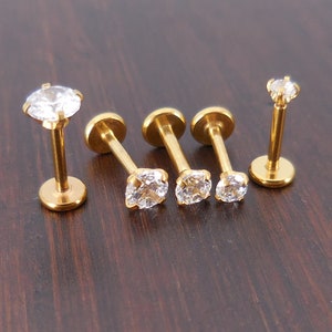 K&ouml;nnte beinhalten: F&uuml;nf goldfarbene Ohr- oder Lippenstecker mit klaren, funkelnden Zirkonia-Steinen. Die Stecker haben eine flache R&uuml;ckseite und einen Stift zum Piercen. Der Schmuck wird auf einer dunkelbraunen Holzoberfl&auml;che pr&auml;sentiert.