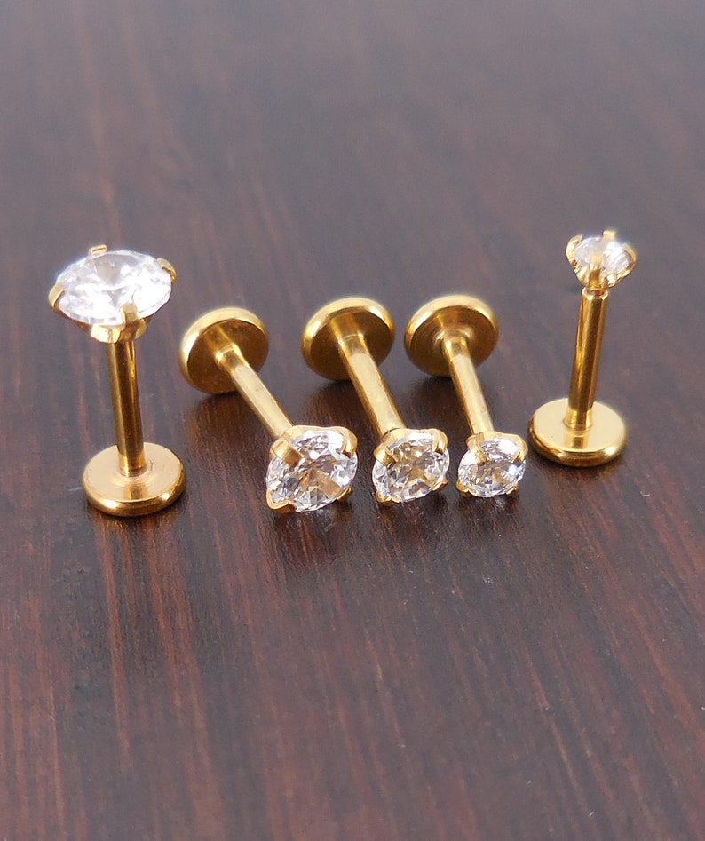 K&ouml;nnte beinhalten: F&uuml;nf goldfarbene Ohr- oder Lippenstecker mit klaren, funkelnden Zirkonia-Steinen. Die Stecker haben eine flache R&uuml;ckseite und einen Stift zum Piercen. Der Schmuck wird auf einer dunkelbraunen Holzoberfl&auml;che pr&auml;sentiert.
