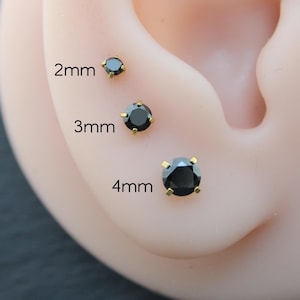 May include: Close-up of an ear with three black stud earrings of varying sizes: 2mm, 3mm, and 4mm. Each earring features a round, black gemstone set in a gold-colored metal. The sizes are labeled in white text.