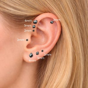 Pode incluir: Close-up de uma orelha com vários brincos de botão pretos. Os brincos são quadrados e variam em tamanho de 2 mm a 4 mm. A orelha é colocada contra um fundo de cabelo loiro.