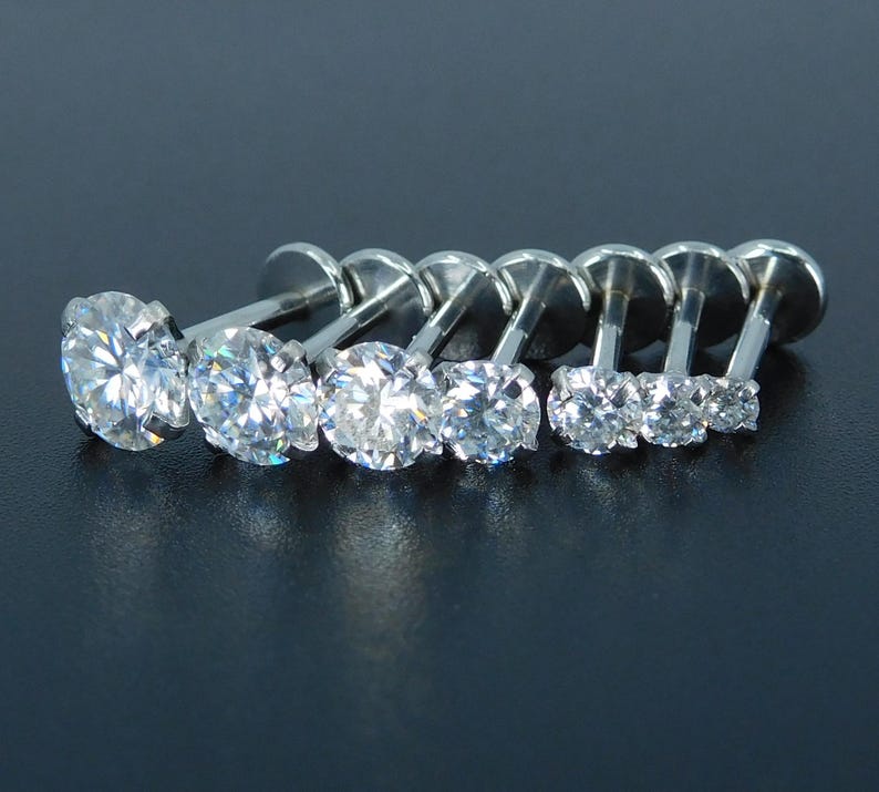 K&ouml;nnte beinhalten: Sieben silberfarbene Labret-Ohrstecker mit klaren, rund geschliffenen Edelsteinen. Die Ohrstecker sind in einer Reihe angeordnet und zeigen einen Gr&ouml;&szlig;enverlauf von gro&szlig; nach klein. Der Schmuck ist vor einem dunklen Hintergrund platziert.