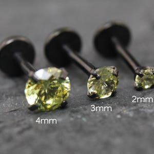 May include: Three black metal labret studs with round, light yellow-green faceted gemstones in four-prong settings. The studs are labelled with sizes 4mm, 3mm, and 2mm, indicating the diameter of the stones. Each piece features a flat back disc.