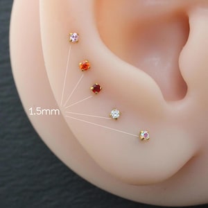 Op de afbeelding: Close-up van een oor met meerdere kleine, kleurrijke oorstekers. De oorbellen zijn goudkleurig en voorzien van gekleurde edelstenen in roze, oranje, rood, wit en iriserende tinten. De tekst "1,5 mm" is zichtbaar.