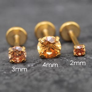 Puede incluir: Tres pendientes de botón con parte posterior plana en tono dorado con piedras preciosas redondas de color naranja en engastes de cuatro garras. Los pendientes están etiquetados con tamaños de 3mm, 4mm y 2mm, indicando el diámetro.