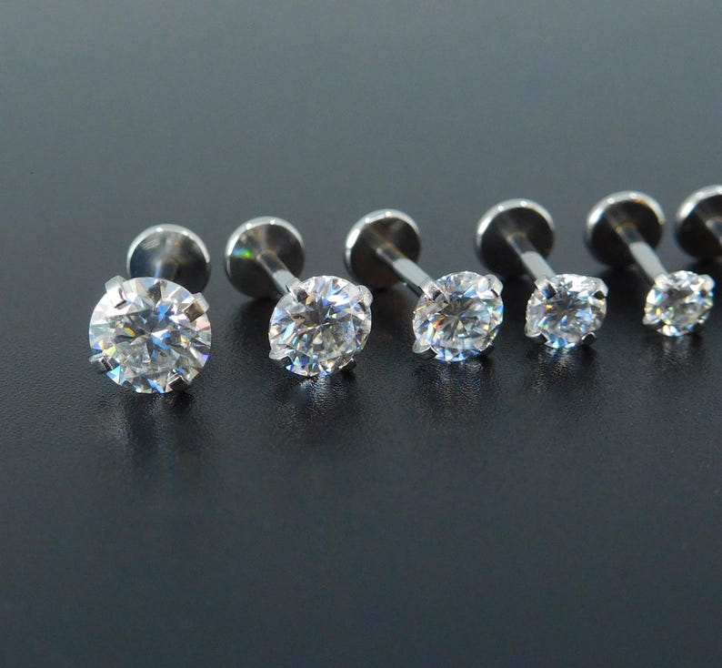 K&ouml;nnte beinhalten: Eine Sammlung von sechs silberfarbenen Labret-Ohrsteckern mit klaren, rund geschliffenen Zirkonia-Steinen. Die Ohrstecker sind in einer Reihe angeordnet und zeigen die unterschiedlichen Gr&ouml;&szlig;en der Edelsteine. Der Schmuck ist vor einem dunklen Hintergrund platziert.