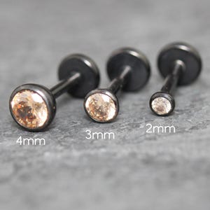 May include: Three black metal labret studs with round champagne-colored gemstones. The studs are arranged by size, labeled 4mm, 3mm, and 2mm, each featuring a flat circular back and a prong-set stone on the front for secure body piercing wear.