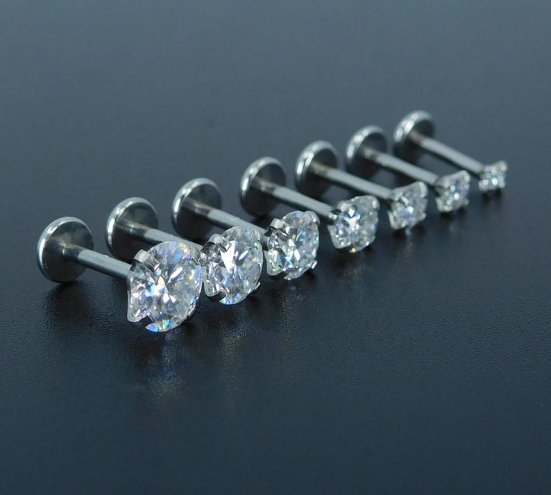 K&ouml;nnte beinhalten: Eine Sammlung von sieben silberfarbenen Lippenpiercing-Steckern mit klaren, funkelnden Zirkonia-Steinen. Die Stecker sind in einer Reihe angeordnet und zeigen verschiedene Gr&ouml;&szlig;en der runden und quadratisch geschliffenen Edelsteine. Der Hintergrund ist dunkel und einfarbig.