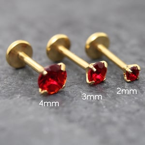 May include: Three gold-colored labret studs with red gemstones. The gemstones are square-cut and vary in size: 4mm, 3mm, and 2mm. The studs have a flat back and a post for insertion.
