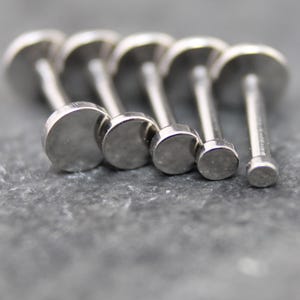 May include: A collection of five silver-toned metal labret studs arranged in a row, each featuring a flat, circular disc top of varying sizes. The studs have a smooth, polished finish and a straight post extending from the back of each disc.