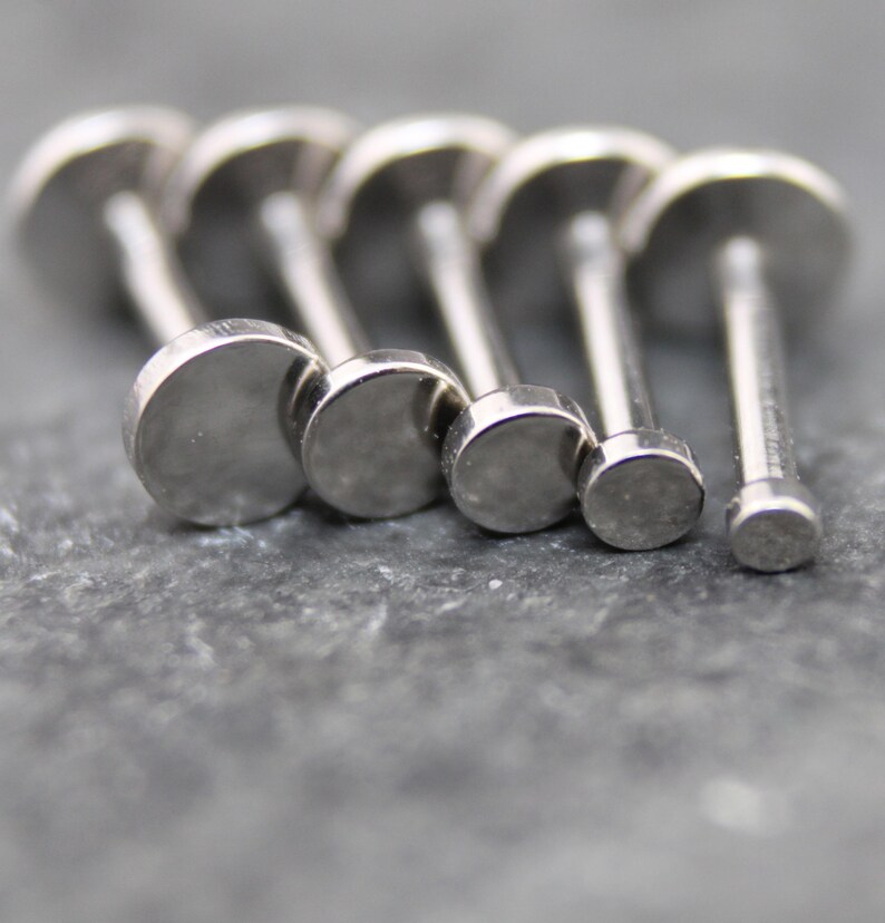 May include: A collection of five silver-toned metal labret studs arranged in a row, each featuring a flat, circular disc top of varying sizes. The studs have a smooth, polished finish and a straight post extending from the back of each disc.