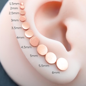 Può includere: Una serie di dieci orecchini a lobo circolari in oro rosa disposti in una linea curva su un orecchio modello. Le dimensioni sono etichettate da 1,5 mm a 6 mm con incrementi di 0,5 mm. Ogni orecchino piatto e rotondo ha una finitura metallica liscia.