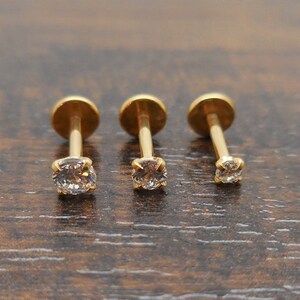 K&ouml;nnte beinhalten: Drei goldfarbene Ohrstecker mit einem kleinen, klaren, runden Edelstein. Die Ohrringe werden auf einer dunkelbraunen Holzoberfl&auml;che pr&auml;sentiert. Die Ohrstecker haben einen Stift und eine runde, flache R&uuml;ckseite.