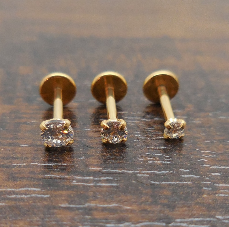 K&ouml;nnte beinhalten: Drei goldfarbene Ohrstecker mit einem kleinen, klaren, runden Edelstein. Die Ohrringe werden auf einer dunkelbraunen Holzoberfl&auml;che pr&auml;sentiert. Die Ohrstecker haben einen Stift und eine runde, flache R&uuml;ckseite.