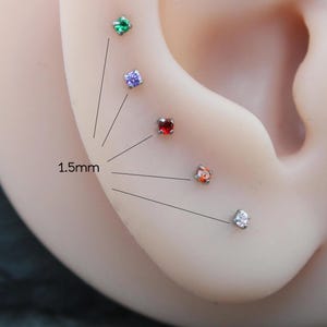 May include: Close-up of an ear with multiple small, colorful gemstone stud earrings. The gemstones are green, purple, red, orange, and clear. The text "1.5mm" is next to the earrings, indicating their size.