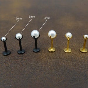 May include: A collection of labret studs with faux pearl tops. The studs vary in size, with pearls measuring 2mm, 3mm, and 4mm. The posts are black or gold-colored, and the pearls are white. The jewelry is displayed on a brown surface.