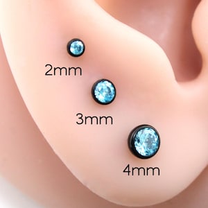 May include: Three round stud earrings with black metal settings and light blue faceted stones. The studs are labeled with their sizes: 2mm, 3mm, and 4mm. Each earring features a circular bezel holding a single sparkling blue gem in place.