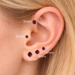 May include: An ear with multiple red square-cut stud earrings in various sizes: 2mm, 3mm, and 4mm. The earrings are set in black metal. The image shows the earrings in place on an ear.