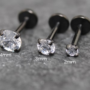 Op de afbeelding: Drie zwarte metalen labret-studs met heldere ronde stenen in een zetting. De studs zijn gelabeld met hun respectievelijke steenmaten van 4mm, 3mm en 2mm. Elk sieraad heeft een platte ronde achterkant en een rechte staaf met een geslepen steen.