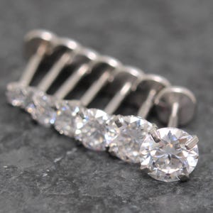 May include: A row of silver-colored stud earrings with clear, round, sparkling gemstones. The earrings are arranged in a diagonal line, with the largest earring in focus at the front. The background is a dark gray, textured surface.