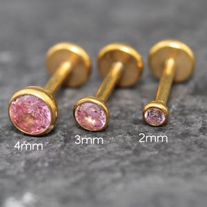 May include: Three gold-tone labret studs with pink circular gemstones in 4mm, 3mm, and 2mm sizes. Each stud features a flat back disc and a bezel-set stone. The text 4mm, 3mm, and 2mm is printed below each corresponding stud to indicate the size.