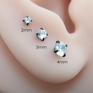 May include: Three round, clear stud earrings set in black metal prongs, displayed in sizes 2mm, 3mm, and 4mm. Each earring features a single faceted stone held by four dark prongs, arranged vertically to show the size difference clearly.
