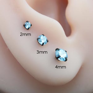 May include: Three round, light blue gemstone stud earrings set in dark metal prongs. The earrings are displayed in ascending sizes labeled 2mm, 3mm, and 4mm, showing the clear, faceted stones held by four-prong settings against a neutral skin tone.