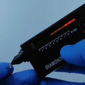K&ouml;nnte beinhalten: Ein schwarzes Diamantpr&uuml;fger&auml;t mit digitaler Anzeige wird verwendet, um einen kleinen, klaren Edelstein zu testen. Das Ger&auml;t hat eine Skala von 1 bis 8 und das Wort "DIAMOND" ist darauf gedruckt. Die Person tr&auml;gt blaue Handschuhe.