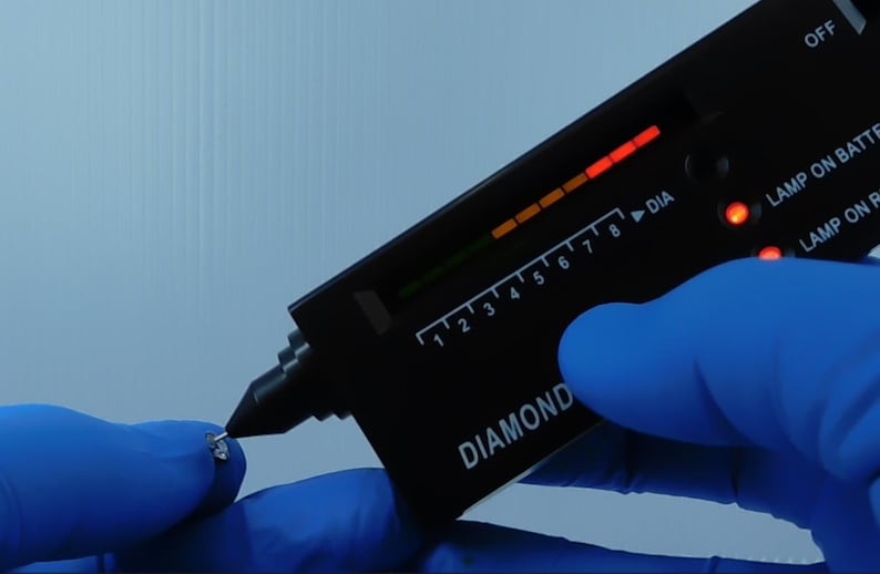 K&ouml;nnte beinhalten: Ein schwarzes Diamantpr&uuml;fger&auml;t mit digitaler Anzeige wird verwendet, um einen kleinen, klaren Edelstein zu testen. Das Ger&auml;t hat eine Skala von 1 bis 8 und das Wort "DIAMOND" ist darauf gedruckt. Die Person tr&auml;gt blaue Handschuhe.