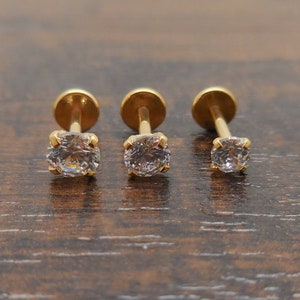 K&ouml;nnte beinhalten: Drei goldfarbene Ohrstecker mit klaren, runden Edelsteinen. Die Ohrringe haben eine vierzackige Fassung und eine flache Scheibenr&uuml;ckseite. Die Ohrstecker werden auf einer dunkelbraunen Holzoberfl&auml;che pr&auml;sentiert.