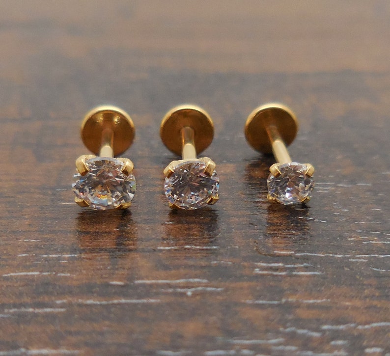 K&ouml;nnte beinhalten: Drei goldfarbene Ohrstecker mit klaren, runden Edelsteinen. Die Ohrringe haben eine vierzackige Fassung und eine flache Scheibenr&uuml;ckseite. Die Ohrstecker werden auf einer dunkelbraunen Holzoberfl&auml;che pr&auml;sentiert.
