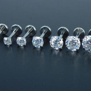 K&ouml;nnte beinhalten: Sieben silberfarbene Labret-Ohrringe mit klaren, runden Zirkonia-Steinen. Die Ohrringe sind in einer Reihe angeordnet, wobei die Steingr&ouml;&szlig;e allm&auml;hlich zunimmt. Die Metallpfosten und -r&uuml;cken sind sichtbar.