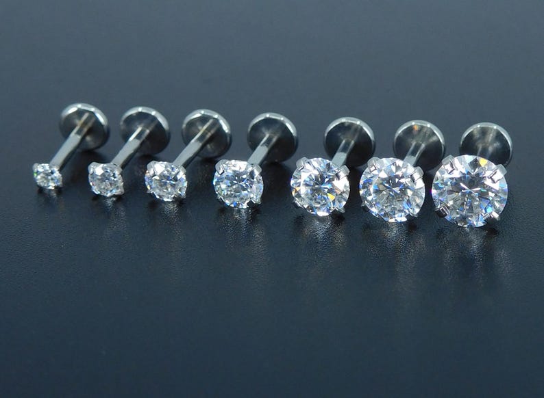 K&ouml;nnte beinhalten: Sieben silberfarbene Labret-Ohrringe mit klaren, runden Zirkonia-Steinen. Die Ohrringe sind in einer Reihe angeordnet, wobei die Steingr&ouml;&szlig;e allm&auml;hlich zunimmt. Die Metallpfosten und -r&uuml;cken sind sichtbar.