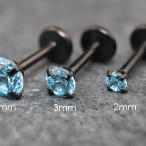 May include: Three black metal labret studs with round, light blue faceted gemstones in four-prong settings. The studs are arranged by size, labeled 4mm, 3mm, and 2mm, each featuring a flat circular back disc and a straight post for body piercing.